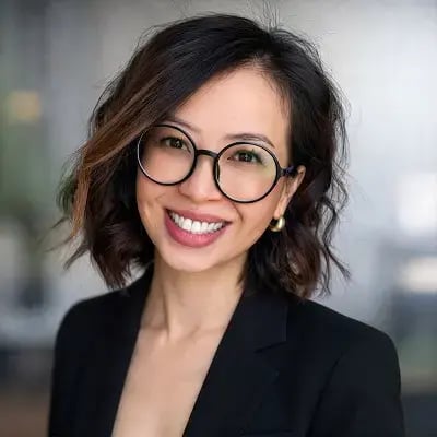 Professional headshot of a smiling person wearing round glasses and a black blazer against a blurred office background.